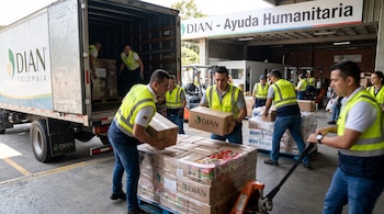Trabajadores con chalecos reflectantes cargan cajas y palés de suministros en un camión con logo DIAN Colombia, frente a un rótulo "DIAN - Ayuda Humanitaria".