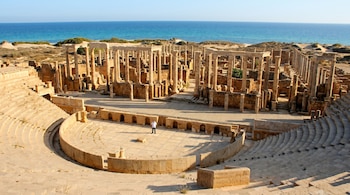 Vista aérea de un teatro romano en ruinas de piedra, con escalones semicirculares y múltiples columnas en la escena, un hombre de pie en el centro y el mar azul de fondo.