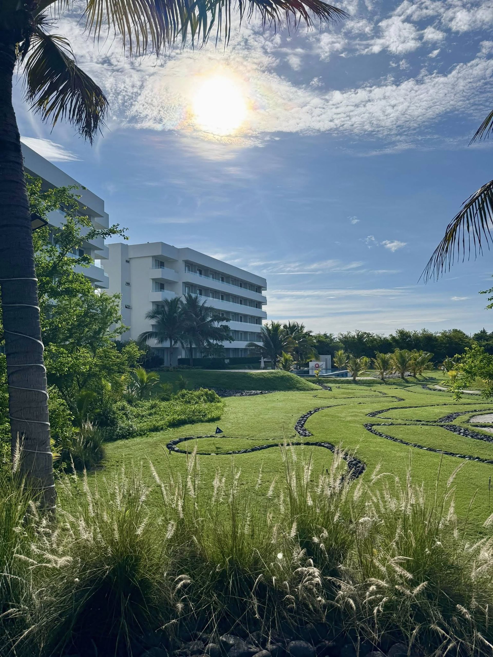 Vista panorámica de un resort moderno con edificios blancos, amplios jardines verdes, palmeras y una piscina bajo un cielo azul brillante con el sol en el centro. ( Oceana Resort-Conventions)