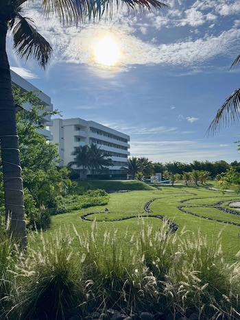 Un paisaje soleado con edificios blancos de un resort, césped verde cuidado con senderos de rocas, palmeras y plantas, bajo un cielo azul claro