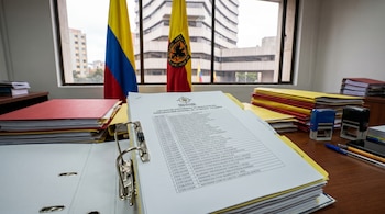 Primer plano de un listado de expedientes de investigación de la Procuraduría General sobre una mesa de madera. Al fondo, banderas de Colombia y edificios.