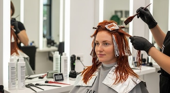 Una mujer de cabello rojizo sentada en una peluquería con papel de aluminio en mechones. Manos con guantes negros aplican tinte con un cepillo. Hay productos y espejos.