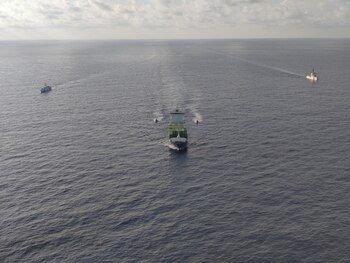 Un buque de carga con contenedores verdes en la cubierta navega en el mar junto a cuatro embarcaciones menores bajo un cielo nublado