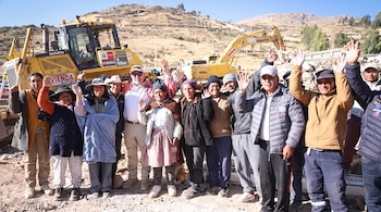 Grupo de personas, incluyendo hombres y mujeres, posa y saluda frente a dos máquinas de construcción amarillas en un terreno árido bajo cielo azul