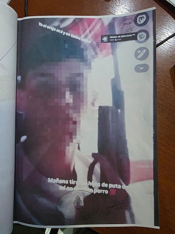 Primer plano de un documento abierto en una mesa de madera que muestra una imagen impresa de un hombre con cara pixelada, un rifle y texto de amenazas. Se ven sellos oficiales