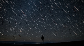 Silueta de una persona de pie en un terreno oscuro, mirando hacia un cielo nocturno repleto de múltiples estelas de luz brillante de meteoros que caen en diagonal.