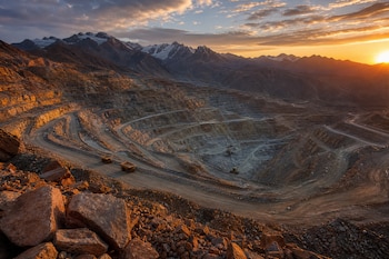 Mina a cielo abierto con terrazas escalonadas, camiones de carga en caminos de tierra y montañas nevadas al atardecer.
