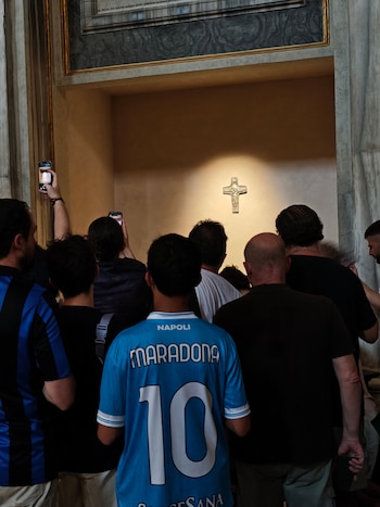 Grupo de personas de espaldas, una lleva camiseta azul con "Maradona 10" y "Napoli". Se observa un crucifijo dorado iluminado en una pared. Dos personas sostienen teléfonos móviles