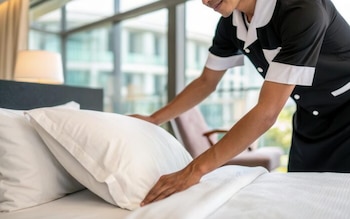 Una persona con uniforme de limpieza de hotel arregla cuidadosamente las almohadas blancas sobre una cama en una habitación luminosa