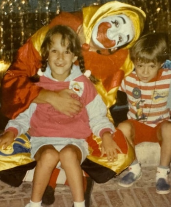 Un payaso con maquillaje blanco, traje rojo y amarillo, abraza a una niña con coletas y un suéter. Un niño con camiseta a rayas los acompaña