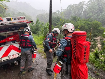 Tres rescatistas con uniformes de la Cruz Roja, cascos y chalecos, junto a un vehículo de rescate blanco. El fondo muestra un paisaje montañoso y húmedo con vegetación