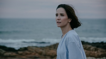 Retrato de Lady M mirando al horizonte, con pelo oscuro al viento. Viste una camisa a rayas azul y blanca, con el mar y rocas al fondo bajo un cielo nublado