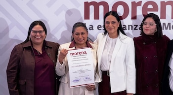 Cuatro mujeres frente a un cartel del partido Morena mientras la mujer del centro sostiene un documento oficial de nombramiento