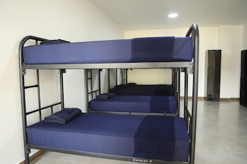 Una fila de tres literas de metal con ropa de cama azul dispuestas en una habitación con paredes claras y taquillas oscuras al fondo