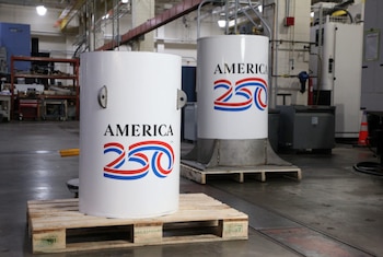 Dos cápsulas cilíndricas blancas con el logo "AMERICA 250" en rojo y azul, apoyadas sobre palés de madera en un entorno industrial