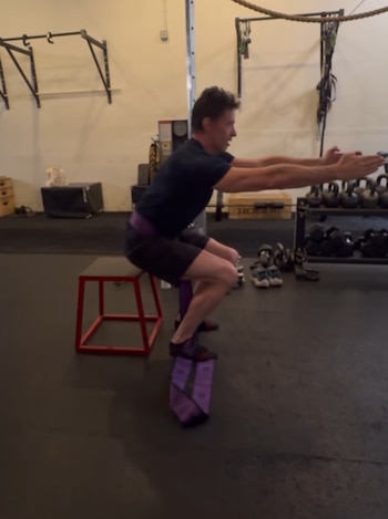 Hombre realiza estocadas con mancuernas en un gimnasio con varias pesas y equipamiento de ejercicio. Varias personas al fondo