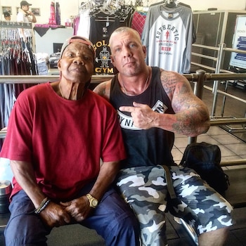 Dos hombres sentados en un banco de gimnasio. Albert Beckles, en camiseta roja y gorra, posa con un hombre joven tatuado que le señala