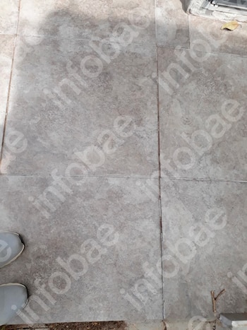 Primer plano de un suelo de baldosas grises con juntas, sombra, parte de dos zapatos, una hoja seca amarilla y una pequeña rama