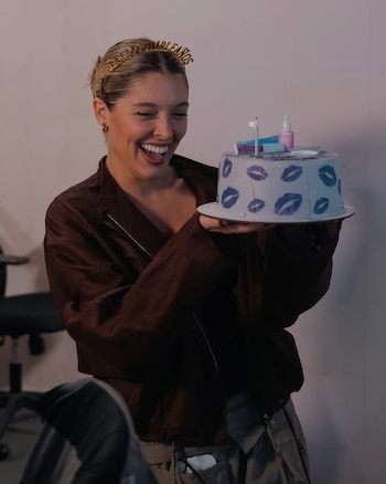 Cami Mayan sonríe sosteniendo un ramo de flores envuelto en papel blanco, vestida con top oscuro, jeans y cinturón dorado, frente a unas escaleras de madera