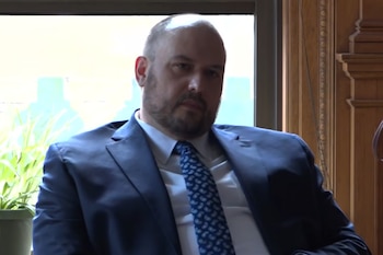 Un hombre con traje oscuro, camisa blanca y corbata estampada azul permanece sentado junto a una ventana