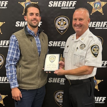Dos hombres, uno con uniforme de sheriff y otro con chaqueta y camisa a cuadros, sostienen una placa conmemorativa frente a logos del sheriff