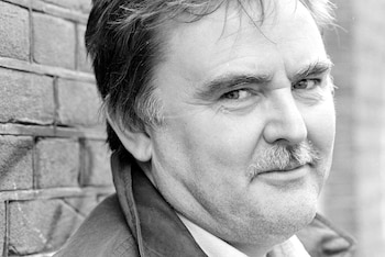 Retrato en blanco y negro de Glen Baxter, un hombre caucásico de mediana edad con bigote, mirando ligeramente a la cámara, con una pared de ladrillo al fondo