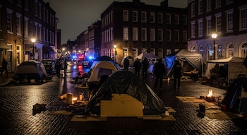 Calle adoquinada nocturna en Savannah, Georgia, con edificios antiguos iluminados, campamentos de tiendas de campaña, fogatas pequeñas y patrullas policiales con luces intermitentes al fondo.