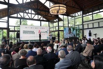 Una audiencia sentada observa el escenario de un evento con dos oradores, una pantalla grande con texto y banderas, y un monitor que muestra a otra persona