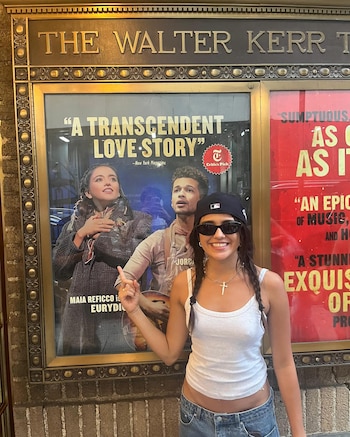 Una mujer joven sonriente con gorra y gafas de sol señala un póster de teatro dentro de un marco dorado, con los nombres 'THE WALTER KERR' arriba