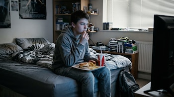 Un joven sentado en la cama sostiene una bandeja con una hamburguesa, papas fritas y un vaso con sorbete frente a un televisor.
