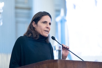Primer plano de una portavoz con cabello castaño oscuro y jersey de cuello alto negro, hablando en un micrófono en un atril de madera con un vaso de agua