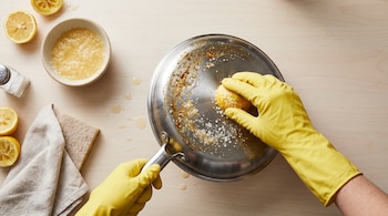 Manos con guantes amarillos sostienen medio limón sobre una sartén de metal, rodeada de rodajas de cítricos, un salero y una esponja