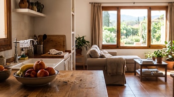 Una mesada de madera con un tazón de fruta, migas de pan y una hormiga, con una cocina y una sala de estar iluminadas de fondo.