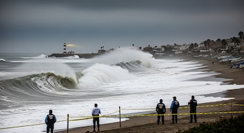 Varios oficiales de Protección Civil observan grandes olas rompiendo en una playa acordonada con cinta amarilla, con un faro y casas costeras al fondo bajo un cielo nublado.