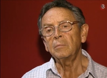 Primer plano de un hombre senior con gafas, cabello oscuro y entrecano, vistiendo una camisa a cuadros clara, mirando al frente sobre un fondo rojo liso