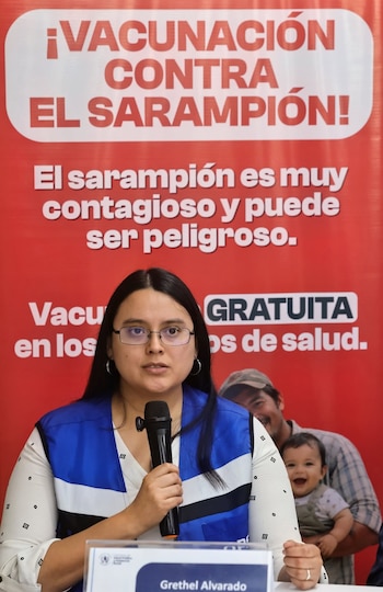 Gretel Alvarado, con gafas y chaleco azul, habla en un micrófono frente a un cartel rojo que anuncia una campaña de vacunación contra el sarampión
