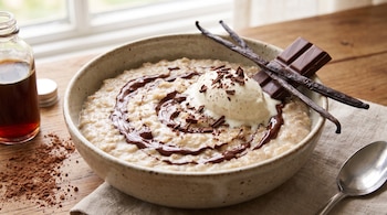 Tazón con avena, jarabe de chocolate, helado, vainas de vainilla y una barra de chocolate junto a una botella y una cuchara sobre una mesa.