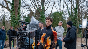 Un hombre con chaqueta llamativa sonríe en primer plano durante un rodaje al aire libre, con cámaras, luces y equipo de producción alrededor.