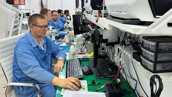 Varios hombres con batas azules trabajan sentados frente a computadoras, cables y componentes electrónicos sobre una mesa de trabajo