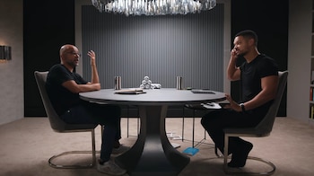 Mo Gawdat y otro hombre sentados frente a una mesa redonda de color gris oscuro en un salón moderno. Mo Gawdat gesticula con su mano derecha mientras habla