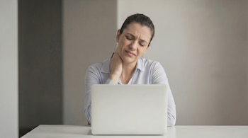 Mujer con camisa a rayas azul y blanca, con los ojos cerrados y la cabeza ligeramente inclinada, sujetándose el cuello con una mano, frente a una laptop blanca.
