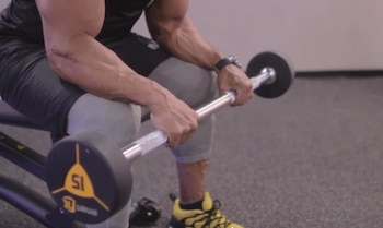 Un hombre en un banco de gimnasio sostiene una barra con pesas, mostrando sus brazos musculosos y un reloj de pulsera