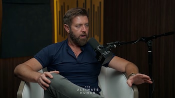 Un hombre con barba viste polo azul y habla frente a un micrófono en un sillón blanco dentro de un estudio