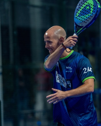 Hombre calvo con camiseta deportiva azul y verde sujeta una raqueta de pádel azul y verde en una cancha. Fondo oscuro