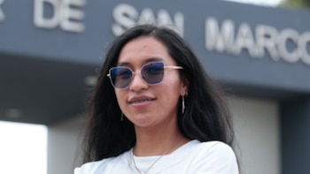 Una mujer joven con lentes oscuros y camiseta blanca posa en primer plano ante un letrero institucional con las palabras San Marcos