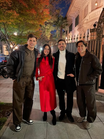 Carolina Baldini, vestida con un abrigo rojo, posa sonriente junto a tres hombres en una calle iluminada de noche, con árboles y un edificio clásico de fondo
