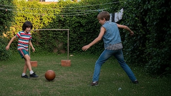 Dos niños juegan al fútbol con un balón de cuero en un patio con césped y una portería improvisada. Se observan tendederos, una parrilla, mesa y sillas.