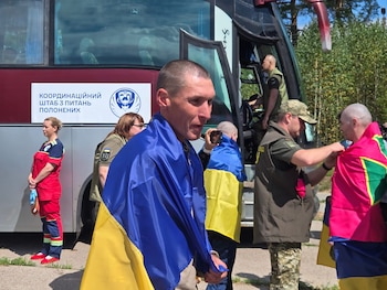 Un hombre con la bandera de Ucrania, otros hombres envueltos en banderas, un militar y una mujer con uniforme sanitario, junto a un autobús en el exterior