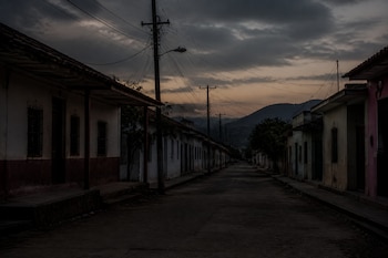 Calle desierta flanqueada por casas bajas y postes de tendido eléctrico frente a montañas bajo un cielo nublado al atardecer.