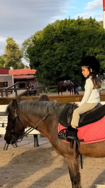 Una niña con casco oscuro y vestimenta de equitación monta un caballo marrón con crin gris y silla con manta rosa, en un picadero de arena